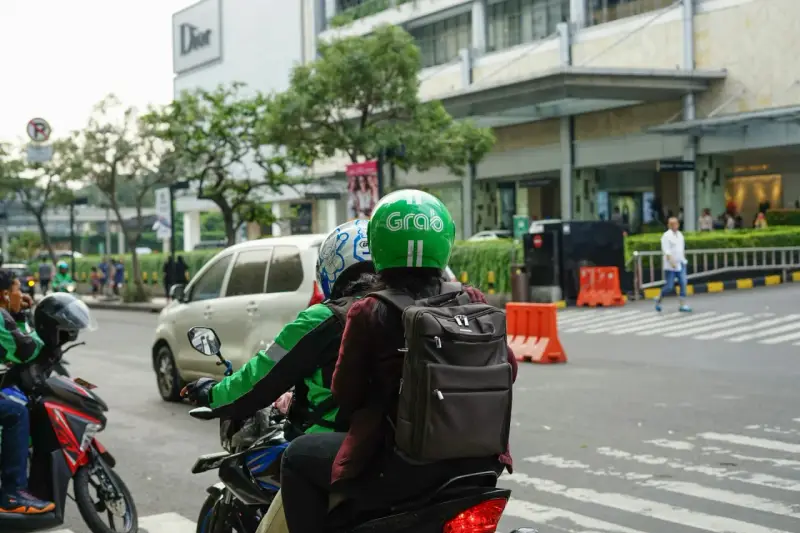 Zwei Personen auf einem Roller, der Fahrer trägt einen grünen Helm mit dem "Grab" Logo. Sie sind unterwegs in Thailand, um mit der Grab App Kunden zu transportieren.