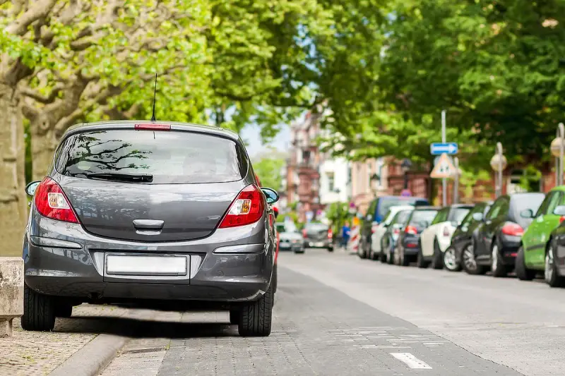 Graues Auto parkt am abgesenkten Bordstein. Mehrere Autos reihen sich die Straße entlang unter grünen Bäumen.